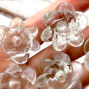 Peut inclure: Une collection de perles en verre transparent et incolore en forme de fleurs &agrave; cinq p&eacute;tales. Chaque perle pr&eacute;sente une surface lisse et arrondie avec un trou central pour l'enfilage, offrant un aspect cristallin avec de fines bulles.
