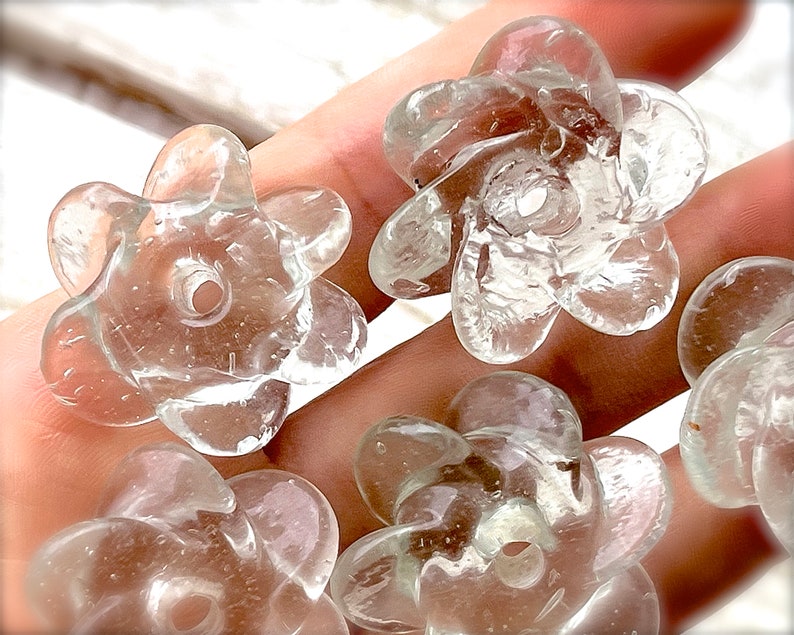 Peut inclure: Une collection de perles en verre transparent et incolore en forme de fleurs &agrave; cinq p&eacute;tales. Chaque perle pr&eacute;sente une surface lisse et arrondie avec un trou central pour l'enfilage, offrant un aspect cristallin avec de fines bulles.