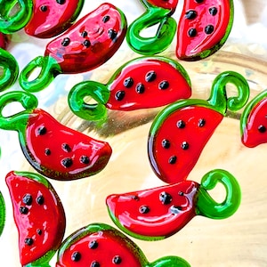 May include: A collection of watermelon-shaped glass charms with green stems. The charms are red and have black seeds.
