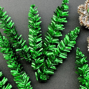 May include: Close-up of shiny green tinsel sprigs and silver and gold star-shaped ornaments with green bead accents. The tinsel has a metallic finish, and the ornaments are detailed with small beads. Festive holiday decorations.