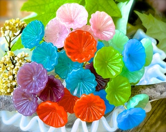 May include: A collection of colorful glass flower-shaped ornaments in shades of blue, green, purple, pink, orange, and red. The ornaments are arranged in a cluster on a white surface.