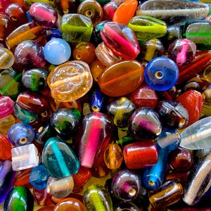 Puede incluir: Una gran colección de cuentas de vidrio variadas en diferentes formas, tamaños y colores, incluyendo rojo, azul, verde, amarillo y morado. Las cuentas presentan acabados lisos, translúcidos y opacos con orificios centrales visibles.