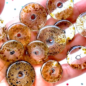 May include: A collection of clear glass beads with a speckled brown pattern and a central hole. The beads are circular and vary slightly in size. The glass has a glossy finish, and the beads are arranged in a close-up shot.