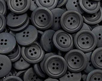 Può includere: Un'immagine ravvicinata di un mucchio di bottoni di plastica neri. I bottoni sono rotondi e hanno quattro fori ciascuno.