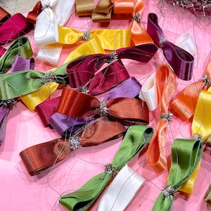 May include: A collection of colorful satin ribbon bows with silver wire accents. The bows are arranged on a pink surface.