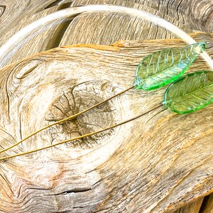 May include: Two green glass leaves on gold wire stems, arranged on a piece of weathered wood.