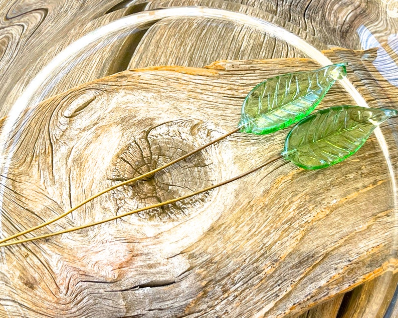 May include: Two green glass leaves on gold wire stems, arranged on a piece of weathered wood.