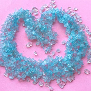 May include: A heart-shaped arrangement of light blue aquamarine chips on a pink background. The chips are scattered around the heart shape, creating a delicate and ethereal look.