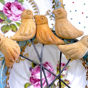 Puede incluir: Cinco pequeñas figuritas de cerámica en forma de pájaro de color marrón sobre tallos de alambre metálico negro. Las figuritas están dispuestas en círculo sobre un plato blanco con un patrón floral azul y dorado.