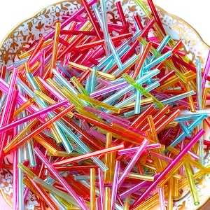 May include: A collection of colorful, translucent glass rods in shades of red, pink, orange, yellow, green, blue, and purple. The rods are scattered inside a decorative, antique-style porcelain dish with a gold rim and floral pattern.