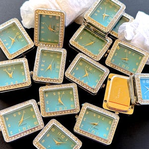 Könnte beinhalten: Eine Sammlung rechteckiger Geneva-Uhren mit türkisfarbenen Zifferblättern und goldenen Zeigern. Die Zifferblätter sind von einem Rand aus kleinen, klaren Kristallen umgeben. Die Uhrengehäuse sind goldfarben. Die Uhren liegen auf einer dunklen Oberfläche.