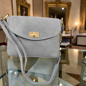 May include: A light blue crossbody bag with a flap closure and gold-tone hardware. The bag features a snakeskin-embossed front and a suede flap. The adjustable strap is also light blue. The bag is rectangular in shape.