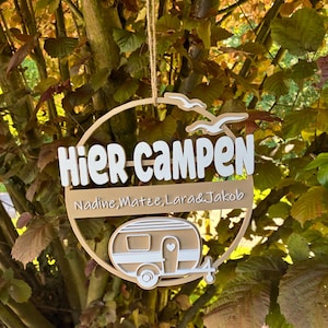 Könnte beinhalten: Runder, beiger Holzschild mit den weißen Worten "Hier Campen", einer Wohnwagen-Illustration und Namen. Zwei weiße Möwen über dem Text. Das Schild hängt an einer natürlichen Schnur. Thema: Camping.