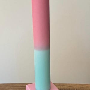 Könnte beinhalten: Eine farbenfrohe Kerze mit einem rosa-türkisen Farbverlauf, die auf einem Stapel geometrischer Formen in Rosa, Gelb und Lila steht. Die Kerze ist ein dekoratives Element.