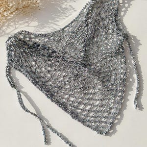 May include: A triangular crochet top featuring a metallic silver open-knit mesh pattern. The garment is constructed with thin, flexible yarn and includes long, slender ties at the corners for fastening, creating a lightweight, semi-sheer appearance.