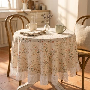 Könnte beinhalten: Ein runder Tisch mit einer floralen Tischdecke und Spitzenbesatz, gedeckt für zwei Personen. Zwei dampfende Tassen stehen auf dem Tisch, zusammen mit einer gefalteten Serviette und einer kleinen Vase mit Lavendel. Zwei Holzstühle umgeben den Tisch.