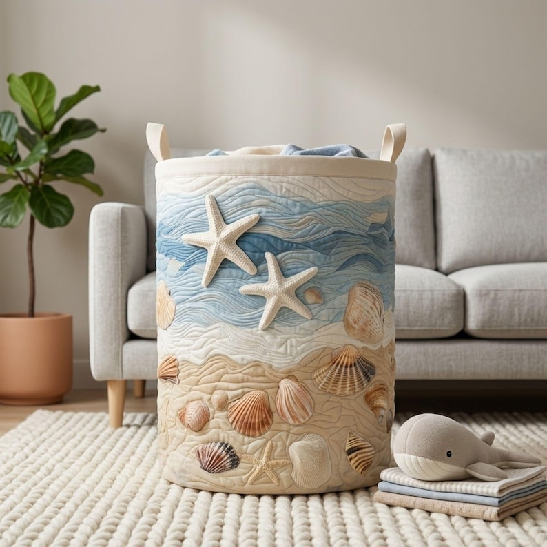 May include: A cylindrical fabric laundry basket featuring a quilted design with a beach theme. The upper section displays blue wavy patterns with two white starfish, while the lower section depicts a sandy shore with various seashells.