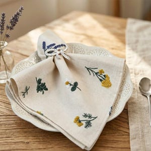 以下が含まれることがあります： ベージュのリネンナプキン。黄色、緑、白の花柄刺繍が施されています。リボン付きのナプキンリングが1枚のナプキンを留めています。ナプキンは、装飾的な縁のある白いお皿の上に置かれ、木製のテーブルに置かれています。