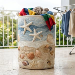 May include: A cylindrical fabric laundry hamper featuring a quilted design with a beach theme. The exterior displays a printed pattern of blue ocean waves, sandy beige shores, two large white starfish, and various seashells in shades of brown.