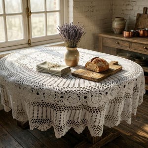 Op de afbeelding: Een witte, ovale gehaakte tafelkleed bedekt een houten tafel. Een vaas met lavendel, een stapel servetten en een houten snijplank met brood staan op de tafel. De scène speelt zich af in een keuken.