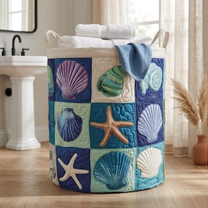 Könnte beinhalten: Ein zylindrischer Wäschekorb aus Stoff mit gestepptem Patchwork-Design, verziert mit applizierten Muscheln und Seesternen in Blau-, Lila-, Grün- und Beigetönen. Der Korb hat zwei Griffe und enthält weiße Handtücher sowie ein blaues Tuch.