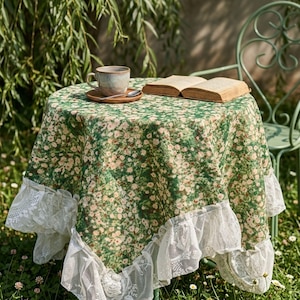Pode incluir: Uma mesa redonda coberta com uma toalha de mesa floral verde, com bordas de renda branca. Uma chávena e pires, juntamente com uma colher, estão sobre a mesa, ao lado de um livro aberto. Uma cadeira de metal verde é visível.