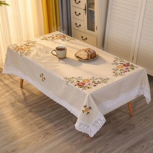 May include: A rectangular beige tablecloth with embroidered floral patterns in shades of orange, pink, and green. The edges are trimmed with white lace. A cup of tea and a plate of bread sit on the table.
