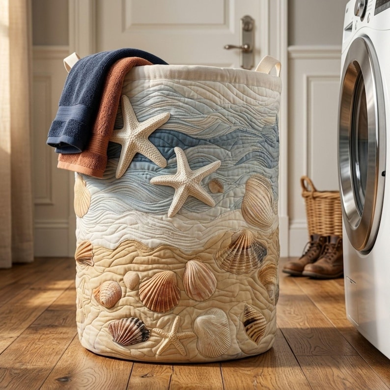 May include: A cylindrical fabric laundry hamper featuring a quilted design with a beach theme. The exterior displays appliqued starfish and various seashells over a textured sand and ocean wave pattern. A navy blue and a rust-coloured towel hang over the rim.