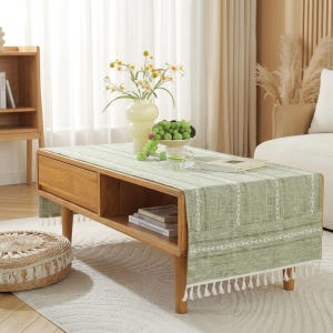 Pode incluir: Uma mesa de centro de madeira castanha clara com um caminho de mesa verde. O caminho de mesa tem um padrão geométrico branco e franjas brancas. Um vaso de flores e uma tigela de uvas estão sobre a mesa. Uma pequena prateleira de madeira está no fundo.