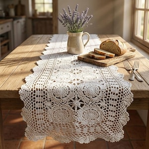 Può includere: Un runner da tavola in uncinetto bianco con un motivo floreale circolare ripetuto e bordi smerlati. Il runner è posizionato su un tavolo in legno insieme a una brocca in ceramica con lavanda, un tagliere in legno, pane affettato e due forchette.