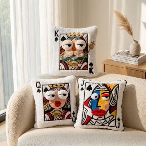 Pode incluir: Três almofadas decorativas de cartas de baralho com bordas brancas e fofas. As almofadas apresentam o Rei, a Rainha e o Valete de Espadas, cada um com um design de rosto único, colorido e estilizado. As almofadas estão em um sofá creme.