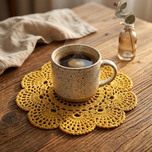 May include: A speckled ceramic mug filled with dark coffee rests on a yellow crocheted coaster with a floral design. A small glass vase with greenery is in the background. The scene is set on a wooden table. The coaster is approximately 15 cm in diameter.