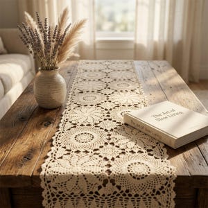 May include: A cream-coloured, crocheted table runner with a floral design on a rustic wooden coffee table. A vase with dried flowers and a book titled "The Art of Slow Living" are also on the table. The scene is illuminated by natural light.