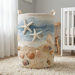May include: A cylindrical fabric storage hamper featuring a quilted design with a beach theme. The upper portion depicts blue and white waves, while the lower portion shows a sandy shore. Appliqu&eacute; starfish and various seashells adorn the exterior.