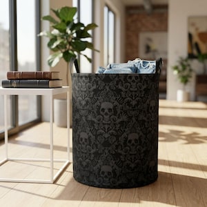 May include: A cylindrical laundry hamper featuring a black and dark grey damask pattern with repeating skull motifs. The hamper has two black handles and is filled with blue denim fabric. The design incorporates intricate scrollwork details.