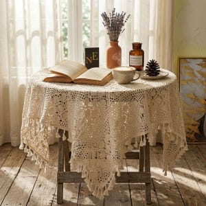 Puede incluir: Un mantel redondo de ganchillo color crema con un ribete de borlas cubre una pequeña mesa de madera. Un libro abierto, una taza de café y un jarrón de lavanda están sobre la mesa. Un pequeño letrero con las letras "NE" también está sobre la mesa.