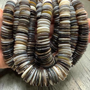 Può includere: Una collana a più fili composta da numerose perline piatte e circolari. Le perline presentano una gamma di colori, tra cui bianco, grigio, nero e marrone. La collana è probabilmente realizzata con materiali naturali e ha un'estetica unica e organica.