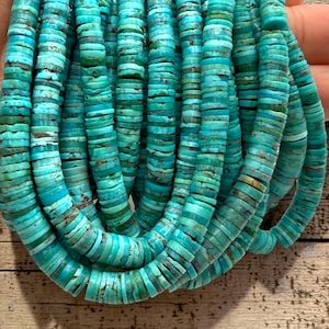 May include: Close-up of strands of turquoise-colored, flat, disc-shaped beads. The beads are a mix of light and dark turquoise, with some showing brown inclusions. The beads are strung together, creating a textured, layered effect. The background is a wooden surface.