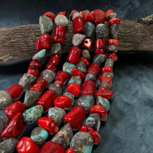 May include: A multi-strand necklace featuring a combination of polished bright red coral beads and irregular, textured turquoise and grey stone beads. The beads are strung together in a series of strands, creating a dense, colorful arrangement.