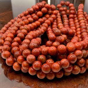 May include: A collection of round, reddish-brown beads strung together. The beads have a speckled appearance with variations in color, ranging from light to dark shades of red and brown. The beads are likely used for jewelry making or crafting.