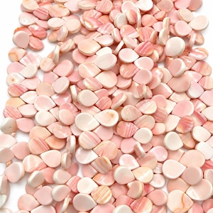 Op de afbeelding: Een grote verzameling kleine, druppelvormige kralen gemaakt van gepolijste roze schelp. Elke kraal heeft een glad, glanzend oppervlak met natuurlijke variaties in lichte en donkere roze tinten, wat de organische, parelmoerachtige textuur van het materiaal benadrukt.