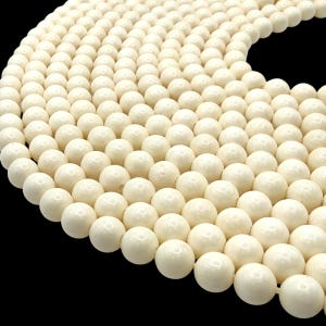 May include: A strand of smooth, round, off-white beads arranged in a curved, semi-circular pattern. Each individual bead features a consistent spherical shape and a uniform, matte-finish cream color, creating a dense, continuous row.