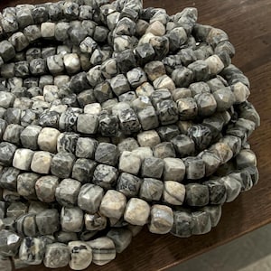 May include: A pile of faceted, square-shaped beads in shades of gray, white, and black. The beads have a marbled appearance, with variations in color and pattern. The beads are strung together, suggesting they are for jewelry making or crafts.