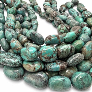 May include: A collection of polished turquoise beads in various shades of blue and green, featuring natural dark brown matrix veining. The beads are smooth, rounded, and irregular in shape, arranged in a dense cluster with a glossy finish.