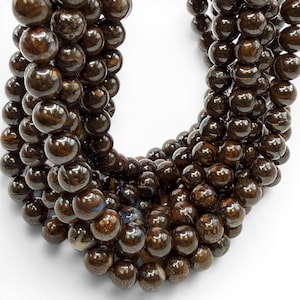 May include: A close-up of a strand of dark brown, glossy beads. The beads are spherical and appear to be made of a polished stone or mineral. The beads are strung together, creating a curved shape against a white background.