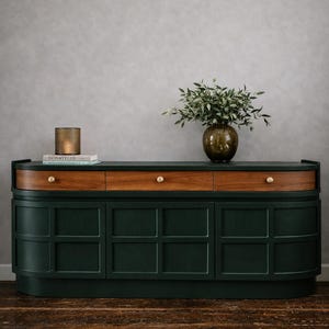 May include: A dark green and wood-toned sideboard with three drawers and cabinet doors. The top features a vase of greenery, a candle, and a book. The cabinet has brass knobs and sits on a wooden floor.