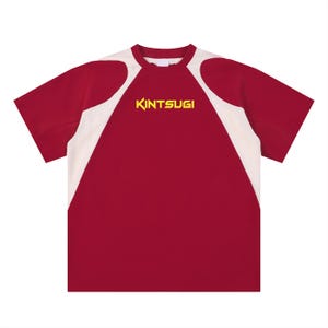 以下が含まれることがあります： 白の肩とサイドパネルが付いた、マルーン色のTシャツ。「KINTSUGI」の文字が胸に黄色でプリントされています。Tシャツはクルーネックで半袖です。