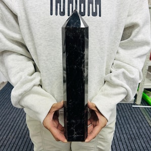 Puede incluir: Una taza de cerámica blanca con asa negra y el texto "But First, Coffee" en negrita negra. La taza se encuentra sobre un fondo borroso, sugiriendo un ambiente acogedor.