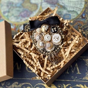 May include: A vintage brooch featuring a collection of gold and silver buttons, clock faces, and a black bow. The brooch has a small crown pendant and is displayed in a brown gift box with shredded paper.