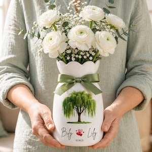 Könnte beinhalten: Eine weiße Keramikvase mit gewelltem Rand, gebunden mit einem grünen Band, gefüllt mit weißen Blumen und Grünzeug. Die Vase zeigt ein Design einer Trauerweide und die Namen "Bily & Lily" mit einem Datum.